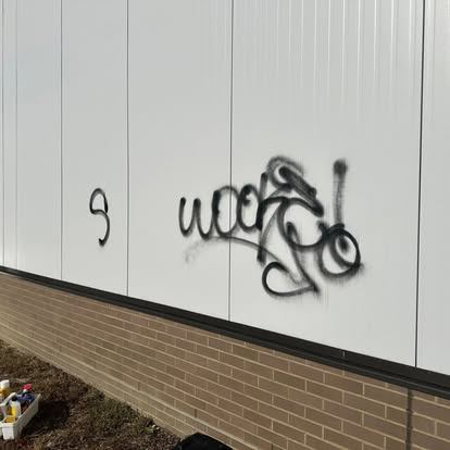 Black spray paint graffiti across a white metal panel wall above a brick base before removal