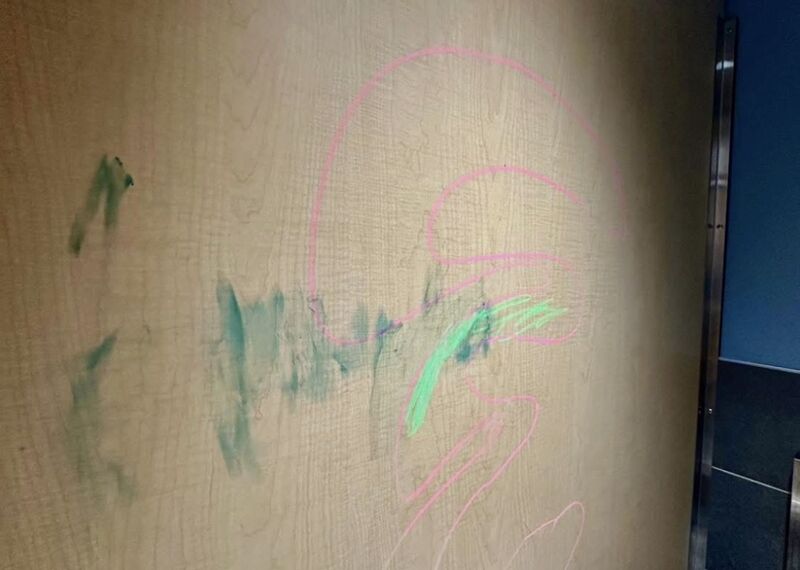 Wood-grain restroom wall panel marked with pink and green marker graffiti and green smudges