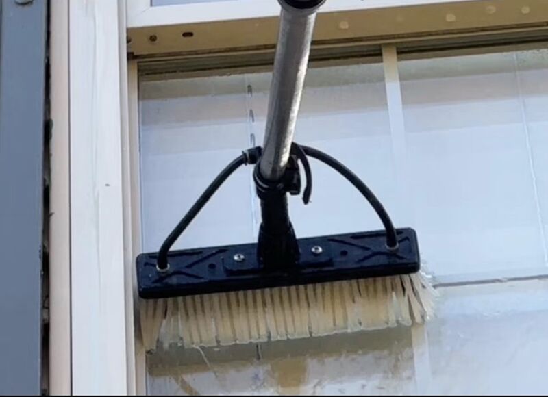 Close-up of a soft-wash brush head on an extension pole rinsing siding beside window trim